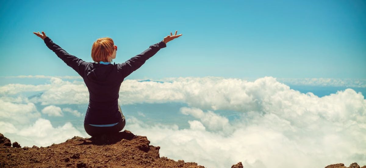 A woman enjoys a scenic view atop a cliff in Kula, Hawaii, surrounded by clouds and blue sky.