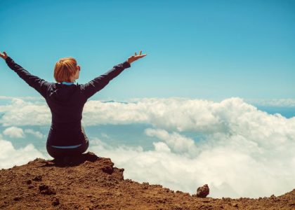 A woman enjoys a scenic view atop a cliff in Kula, Hawaii, surrounded by clouds and blue sky.